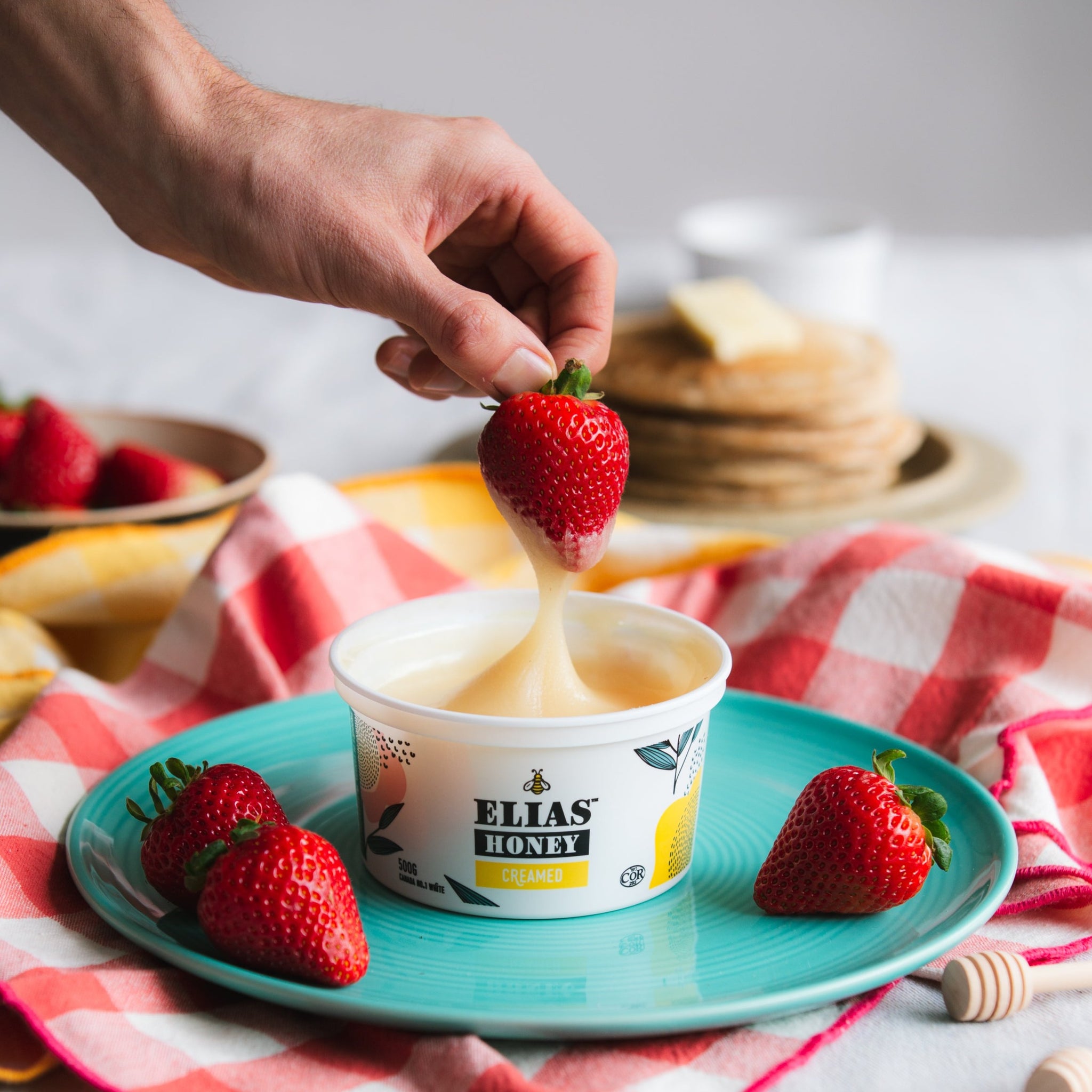 A hand dipping a strawberry in a Elias Honey Creamed 500gr tub