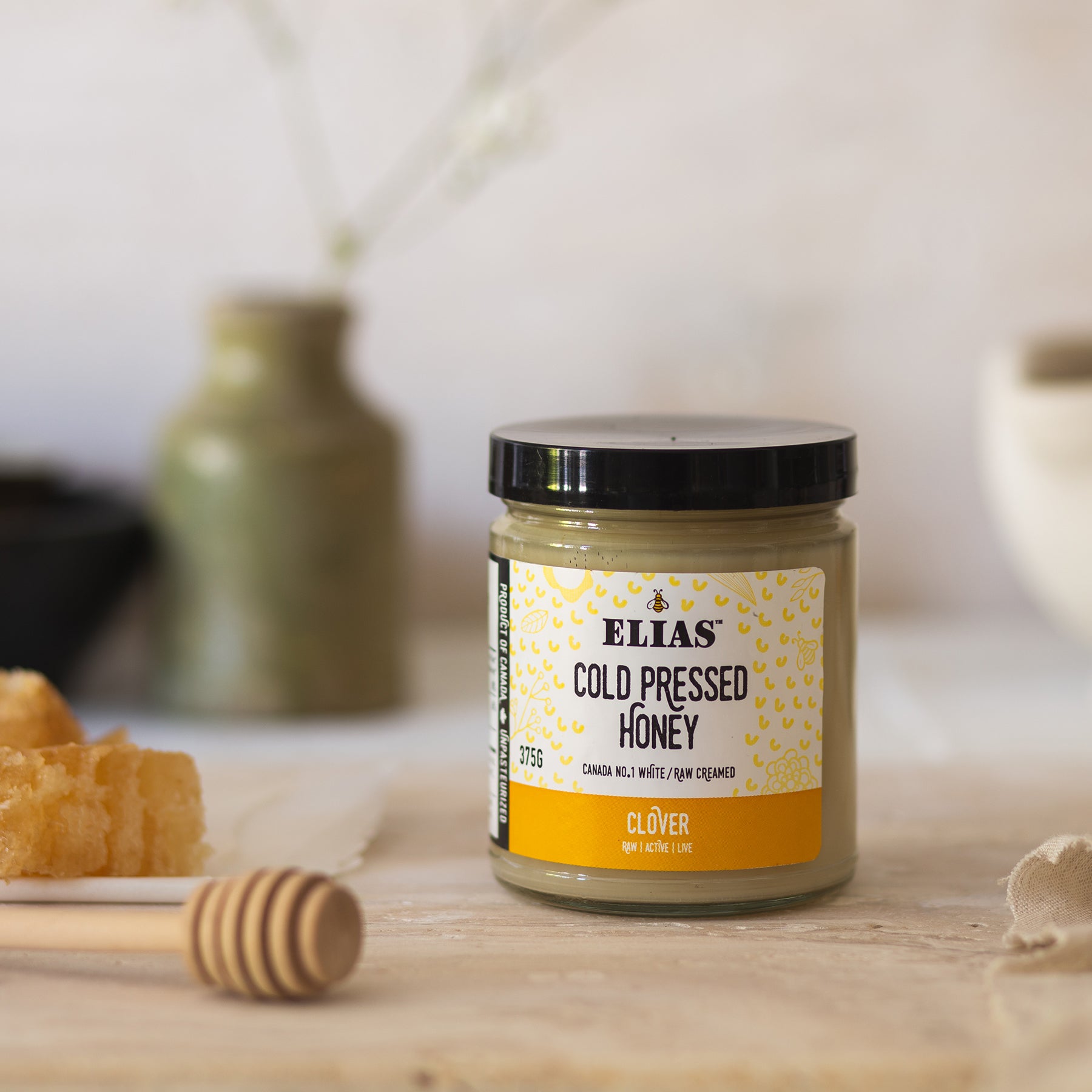 Image of Elias Canadian Cold Pressed Clover Honey in front of a vase and flowers and beside honeycomb.