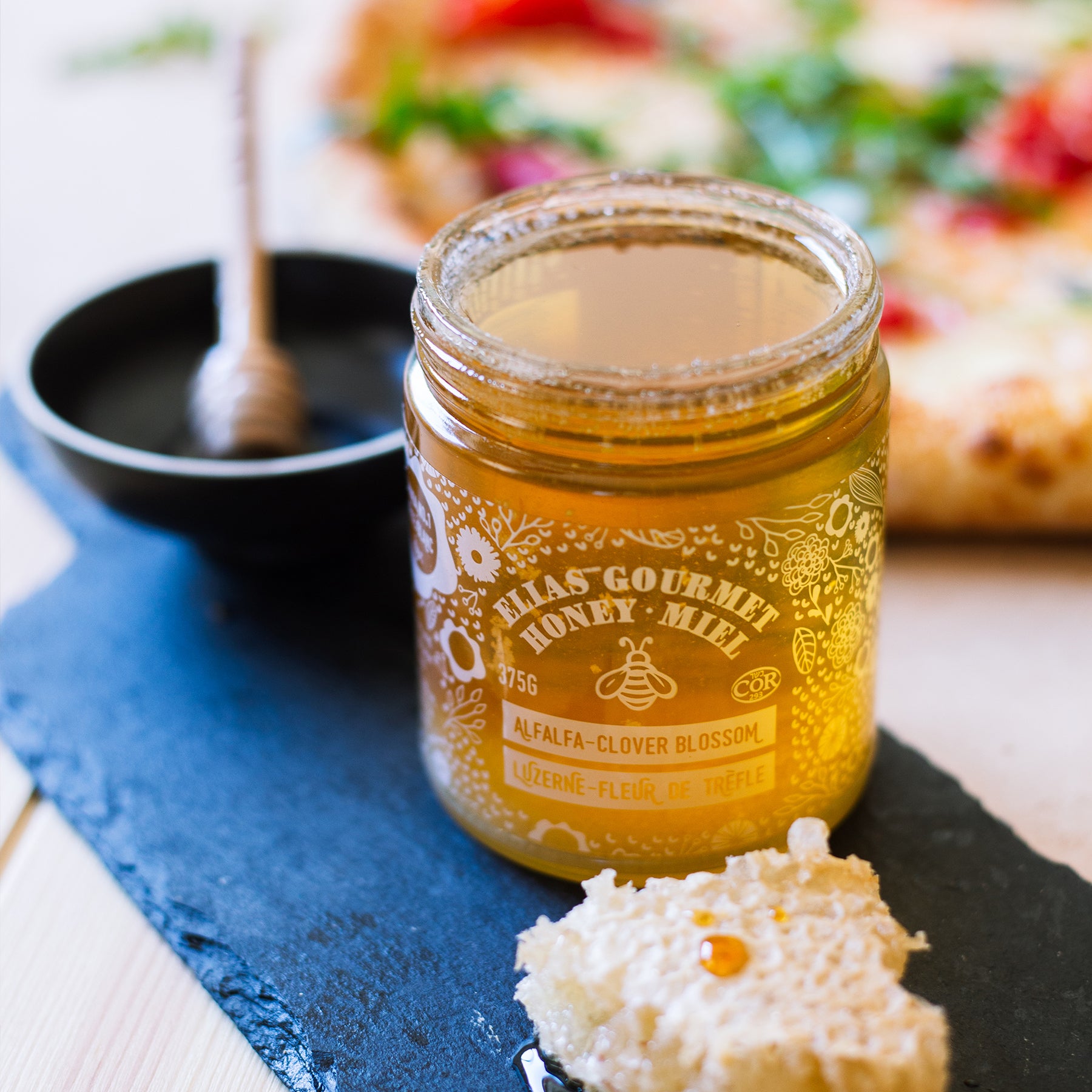Image of Elias Gourmet Raw Alfalfa-Clover Blossom Honey in front of pizza and honey stick in bowl