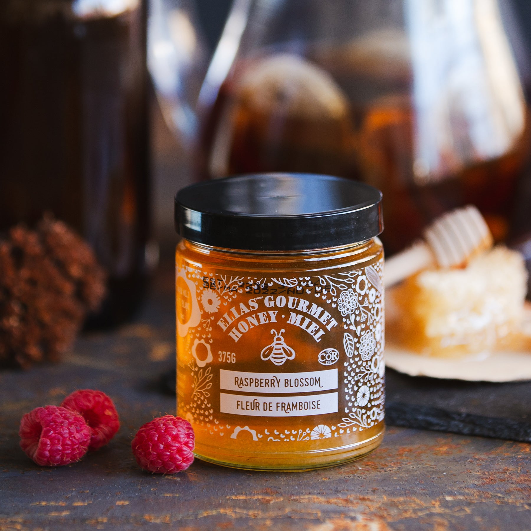 Elias Gourmet Raspberry Blossom Honey Jar with raspberries and honey stick in honeycomb in the background.
